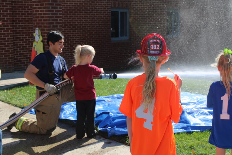 A firefighter kneels on the ground, helping a young girl spray water from a fire hose. Two other children in sports shirts watch nearby, standing on grass beside a brick building.