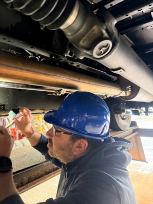 A mechanic wearing a blue hard hat and glasses inspects the undercarriage of a vehicle.